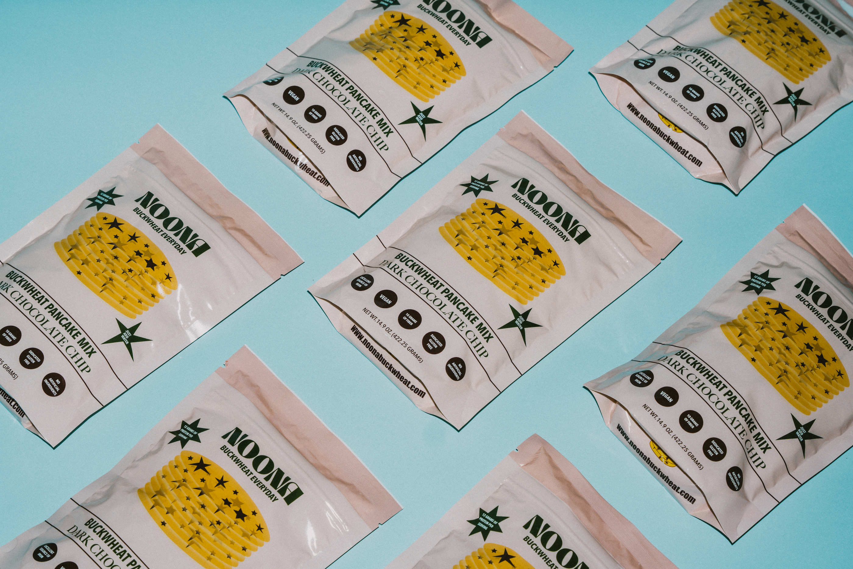 Seven Noona Dark Chocolate Chip Buckwheat Pancake mix packets are lying flat on a baby blue background, with the front of the packaging visible, showcasing the Noona branding and design.