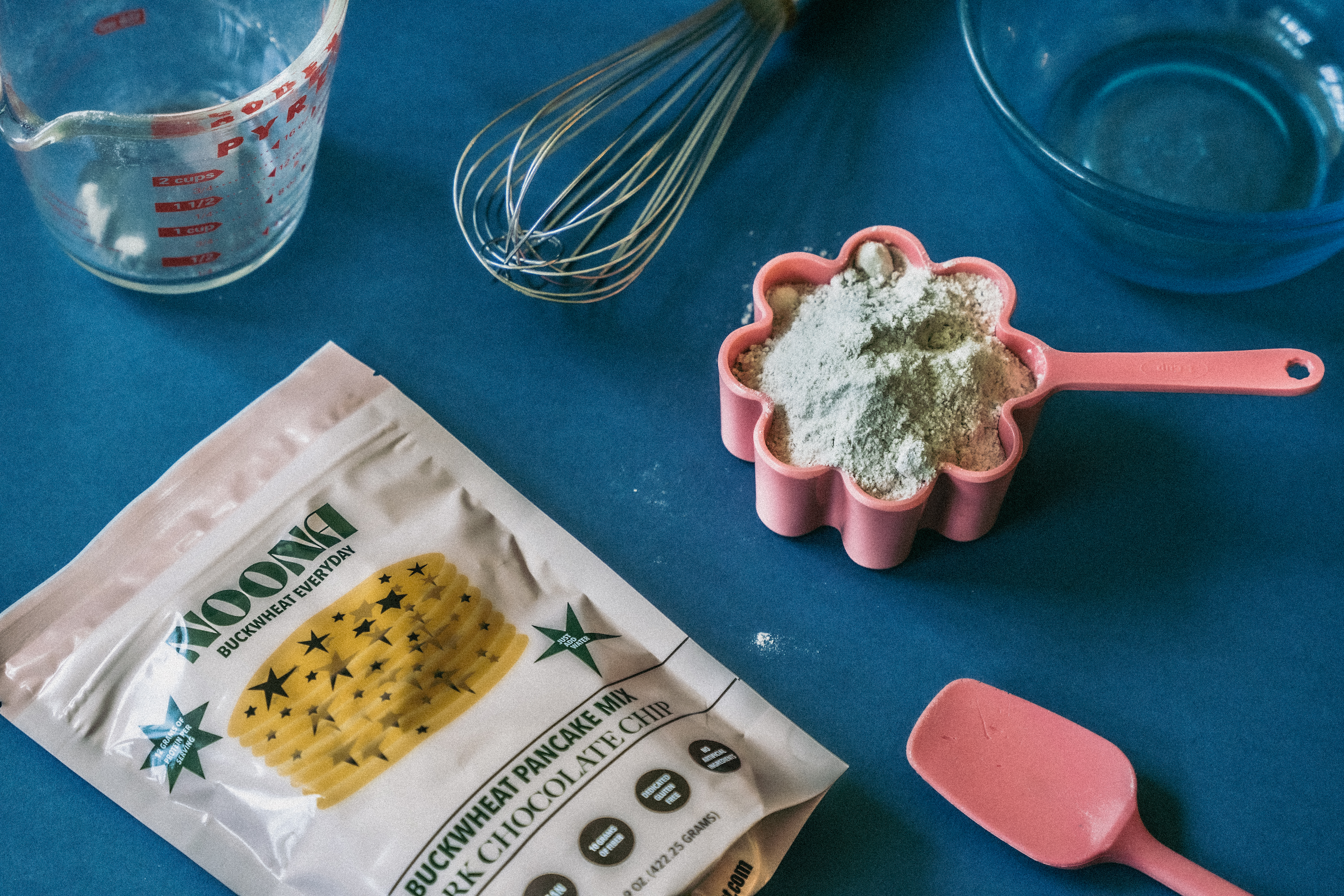 A flat lay of baking tools and ingredients for making Noona buckwheat pancakes on a blue background. The scene includes a bag of Noona Dark Chocolate Chip Buckwheat Pancake Mix, a measuring cup filled with pancake mix, a whisk, a glass bowl, a spatula, and a glass measuring cup.