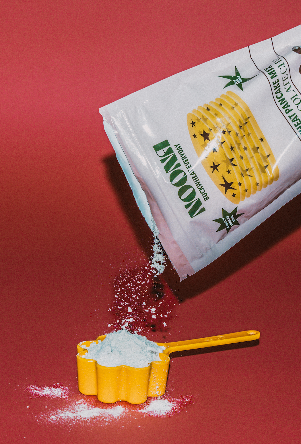 A bag of Noona Buckwheat Pancake Mix is being poured into a bright yellow, flower-shaped measuring cup, with some mix spilling onto a vibrant red background. The Noona packaging is visible, showcasing the branding and design. The image highlights the process of preparing the pancake mix.